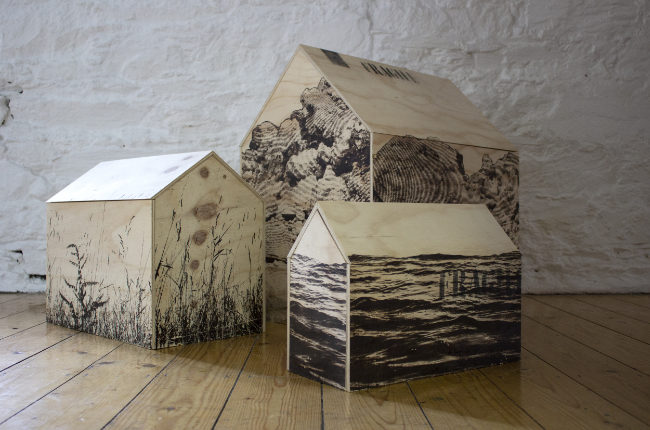Three wooden house-shaped objects with black-and-white nature imagery sit on a wooden floor against a white brick wall.